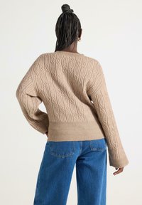 Pullover beige lavorato a maglia con motivo a trecce, maniche ampie e orlo a coste. Indossato con jeans blu a vita alta, visto da dietro.