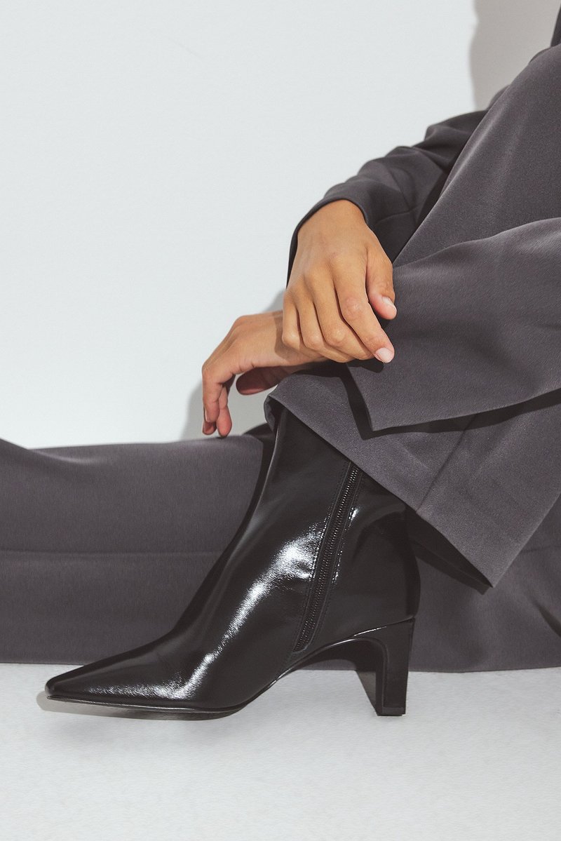 Botines de charol negro con puntera afilada y un tacón cuadrado de altura media. Presentan un zipper lateral y una textura suave y brillante.