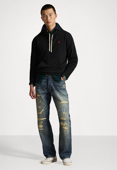 Svart hettegenser med kengurulomme, lagd over en grønn rutete skjorte, kombinert med slitt blå jeans. Skoene er lyse, slip-on stil.