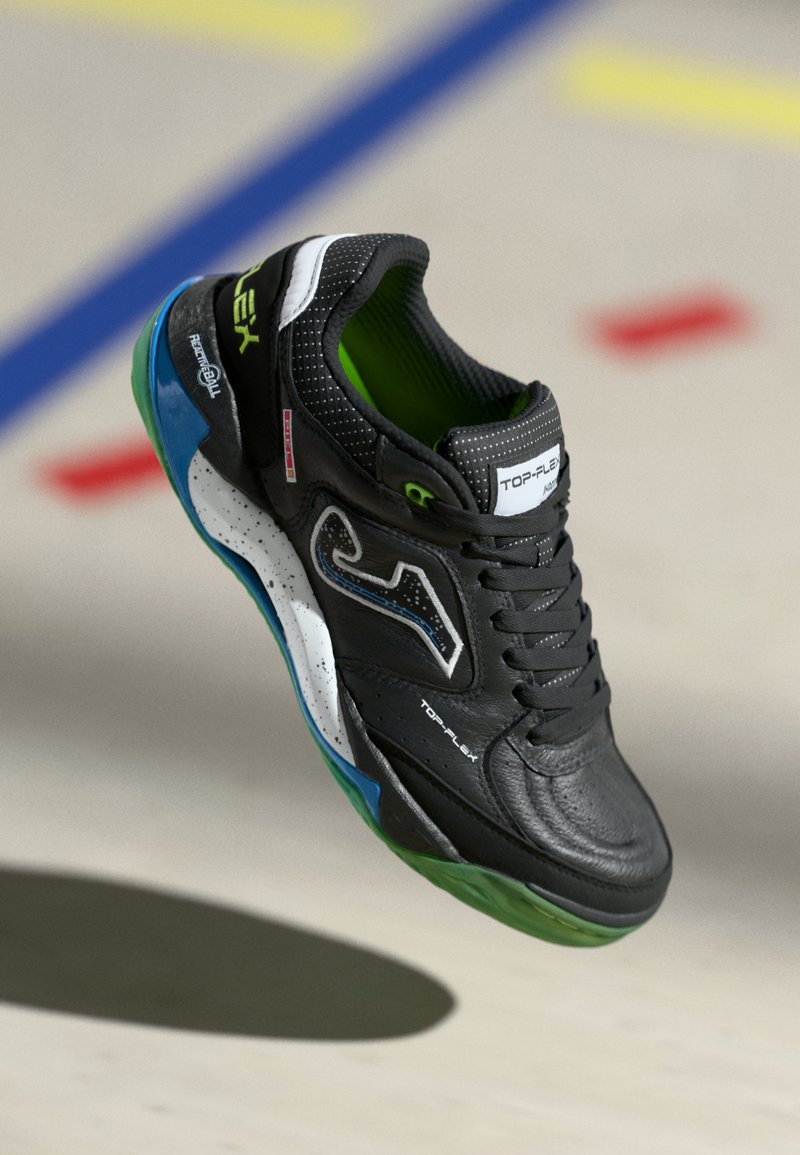 Chaussure de sport noire avec des accents verts, tige en cuir synthétique, texture perforée, semelle blanche avec des détails bleus et verts, et un design épuré.