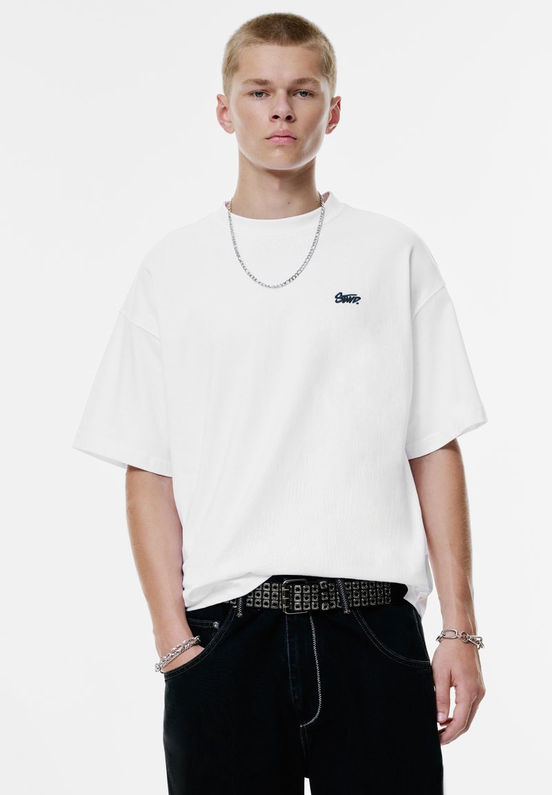 Hvid oversized t-shirt lavet af tekstureret bomuld, med et lille sort logo på brystet. Kombineret med sorte jeans, nittebælte og sølvkæde.