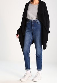 Un cardigan en maille noir sur un t-shirt gris, associé à un jean bleu taille haute. Comprend des poignets retroussés et des baskets blanches. Look simple et décontracté.