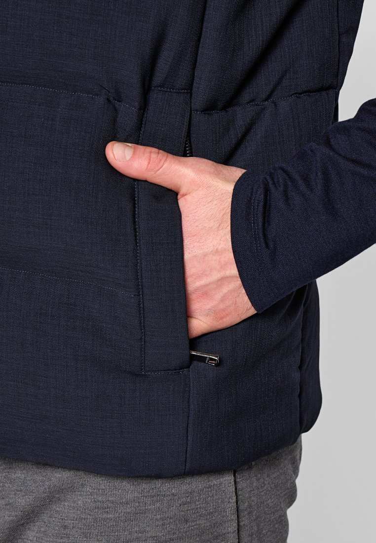 Marinejacke aus strukturiertem Material, mit einem Reißverschluss-Seitenfach und taillierter Passform. Eine Hand befindet sich zur Perspektive in der Tasche.