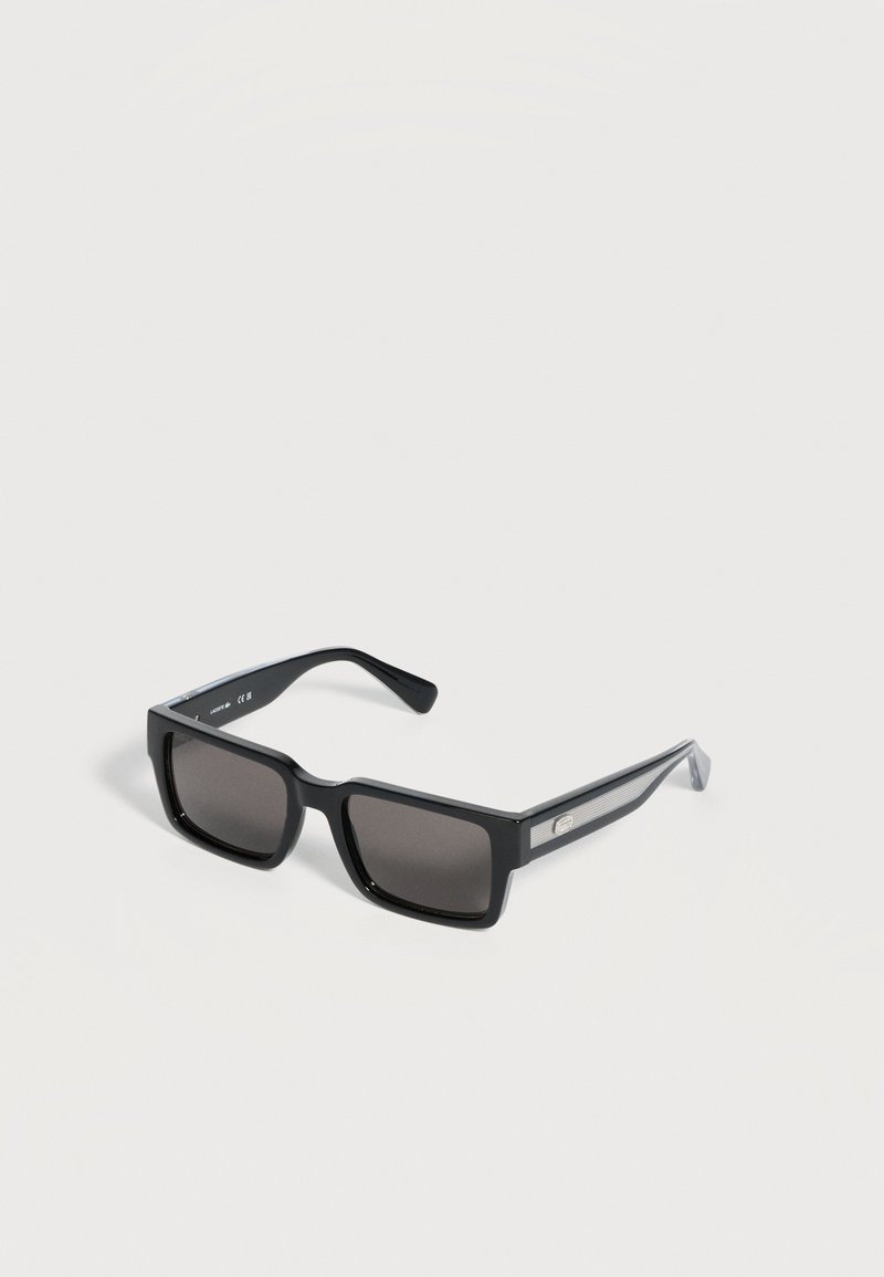 Lunettes de soleil rectangulaires noires avec des verres teintés foncés et des branches texturées avec un petit logo métallique sur le côté.