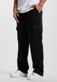 Pantaloni de trening negri, tip cargo, cu două buzunare mari pe părțile laterale, textură răsucită și o croială lejeră, asortați cu adidași albi. Design simplu și practic.