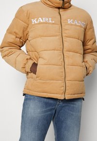 Persona che indossa una giacca puffer di velluto a coste color beige con la scritta "KARL KANI" sul petto, con le mani in tasca, abbinata a jeans di denim blu.