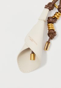 Valge nahast võtmehoidja sujuva pinnaga, millel on pruun nöör ja kuldsetes tooni aktsendid. Sisaldab ümarat, volditud kujundust.