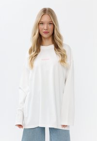 Oversized witte longsleeve shirt met een subtiele roze "MIRONS" print op de voorkant. Zacht materiaal, comfortabele pasvorm en geribbelde halslijn.