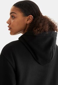 Juoda gobtuvu džemperis su minkšta tekstūra, matomomis siūlėmis ir laisvu kirpimu; dėvima su plaukais atitrauktais nuo veido, rodant auskarą.