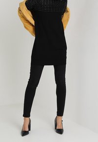 Personne portant une robe noire cloutée, des leggings noirs, des talons hauts noirs, et tenant une veste jaune moutarde derrière son dos.