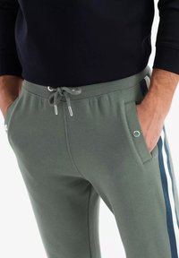 Khaki-Sweatpants mit einem Kordelzugbund, Seitentaschen und blauen sowie weißen Streifenakzenten entlang der Beine. Weicher, strukturierter Stoff.