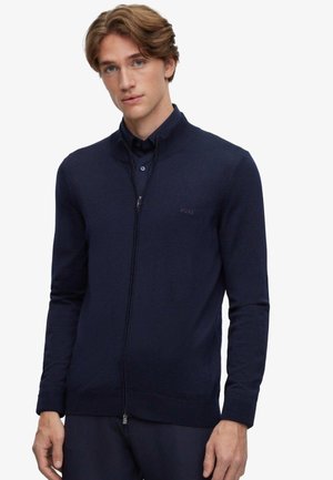 Jeune homme aux cheveux châtain clair portant un pull zippé bleu marine par-dessus une chemise boutonnée, debout devant un fond clair uni.