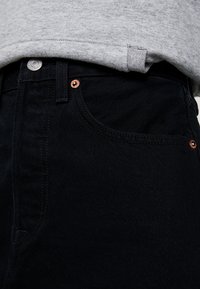 Vaqueros de mezclilla negra con un corte clásico, que presentan remaches de cobre, costuras detalladas y un ligero acabado texturizado. Combinados con una sudadera gris.
