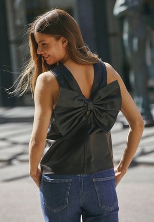 Top in pelle nera con un grande fiocco decorativo sul retro, abbinato a jeans in denim blu. Texture liscia e design senza maniche.