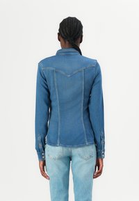 Giubbotto in denim blu scuro con un design aderente, dettaglio della pattina appuntita e polsini con bottoni. Indossato sopra jeans blu chiaro.