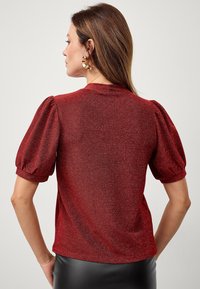 Rotes, strukturiertes Oberteil mit bauschigen kurzen Ärmeln und rundem Ausschnitt; hat einen subtilen Glanz; kombiniert mit einem schwarzen Lederrock.