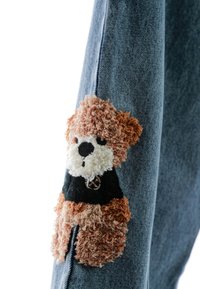 Tejido de mezclilla en azul claro presenta un bordado texturizado y suave de un oso de peluche en marrón y crema, que lleva una camisa negra con un emblema.