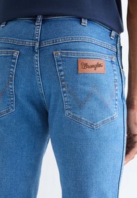 Artimo vaizdas žmogaus, dėvinčio mėlynas Wrangler džinsus, rodančius užpakalines kišenes, siūles ir odinę prekės ženklo detalę dešinėje kišenėje.