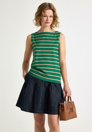 Giovane donna che indossa un top smanicato a righe verdi e beige e una gonna di jeans scuro, tenendo in mano una piccola borsa marrone.