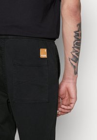 Schwarze Cargo-Hosen mit einer Gesäßtasche und einem quadratischen braunen Etikett. Der Stoff scheint strapazierfähig mit einer glatten Textur zu sein. Einfaches Design mit funktionalen Details.