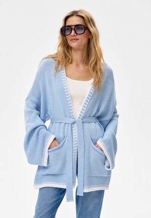 Kobieta ubrana w oversize'owy jasnoniebieski dziergany kardigan z białą lamówką i paskiem, biały tank top, niebieskie dżinsy oraz duże kwadratowe okulary przeciwsłoneczne.