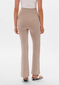 Beżowe spodnie z wysokim stanem w fasonie dzwonów, wykonane z elastycznego materiału. Gładka tekstura, pełna długość, wykończone brzegi, w połączeniu z białymi sandałami na obcasie.