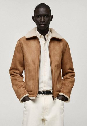 Veste en suède artificiel marron avec un col en fausse fourrure crème, fermeture zip et poches latérales. Portée par-dessus une chemise blanche, associée à un pantalon de couleur claire.