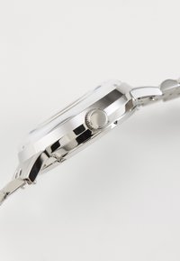 Edelstahluhr mit einem abgerundeten silbernen Gehäuse, Krone an der Seite und einem verbundenen Metallarmband; reflektierende Oberfläche und polierte Verarbeitung.