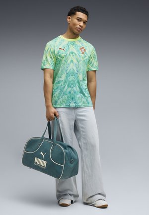Jeune homme portant un maillot de football portugais vert à motif, un jean gris clair à jambes larges, des baskets blanches, tenant un sac de sport Puma turquoise.
