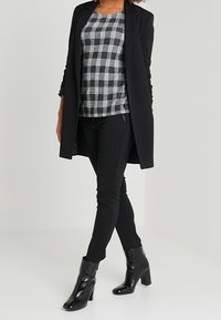 Femme portant un blazer noir, un pantalon ajusté noir, un haut à carreaux gris et noir, et des bottines noires brillantes, se tenant jambes croisées.