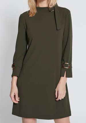 Vrouw die een olijfgroene jurk met lange mouwen draagt, voorzien van gespsieringen op de halslijn en manchetten, staand tegen een eenvoudige lichtgrijze achtergrond.