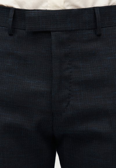 Κοντινή εικόνα σε σκούρο υφασμάτινο παντελόνι με υφή, με θηλιές για ζώνη και κρυφό φερμουάρ, που ξεκουράζεται σε λευκό πουκάμισο.