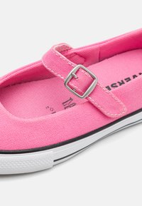 Roze platnena cipela s Mary Jane remenom, koja ima srebrnu metalnu kopču, bijeli gumeni potplat i crni akcent duž ruba.
