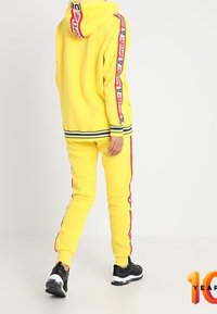 Geel hoodie en bijpassende joggingbroek van zachte stof, met rode zijkanten met logo, zwarte accenten en geribbelde manchetten.