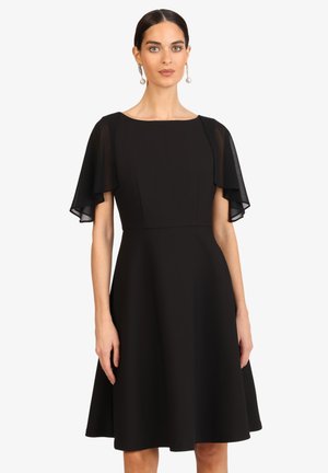 Frau trägt ein schwarzes knielanges Kleid mit transparenten Flatterärmeln und steht vor einem weißen Hintergrund.