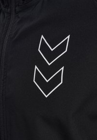 Chaqueta de tela negra con un logotipo geométrico en forma de chevrón blanco en el lado izquierdo. Textura suave con cierre de cremallera en el frente.