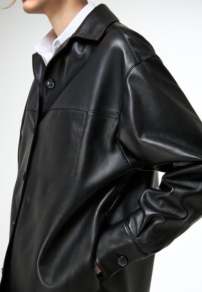 Közeli kép egy személyről, aki fekete bőrgombos kabátot visel mellkasi zsebbel, látható fehér inggallér, keze az oldalzsebben.