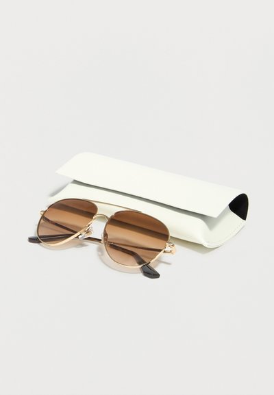 Καφέ-χρωματιστά γυαλιά ηλίου αεροπόρου με χρυσά σκελετούς τοποθετημένα μπροστά από μία λευκή ορθογώνια σκληρή θήκη σε ανοιχτόχρωμη επιφάνεια.