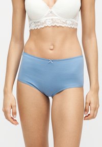 Γυναίκα φοράει λευκό δαντελωτό σουτιέν bralette και γαλάζιο βαμβακερό εσώρουχο με ψηλή μέση και μικρή διακοσμητική φιόγκο, στέκεται με χαλαρά χέρια δίπλα στο σώμα.