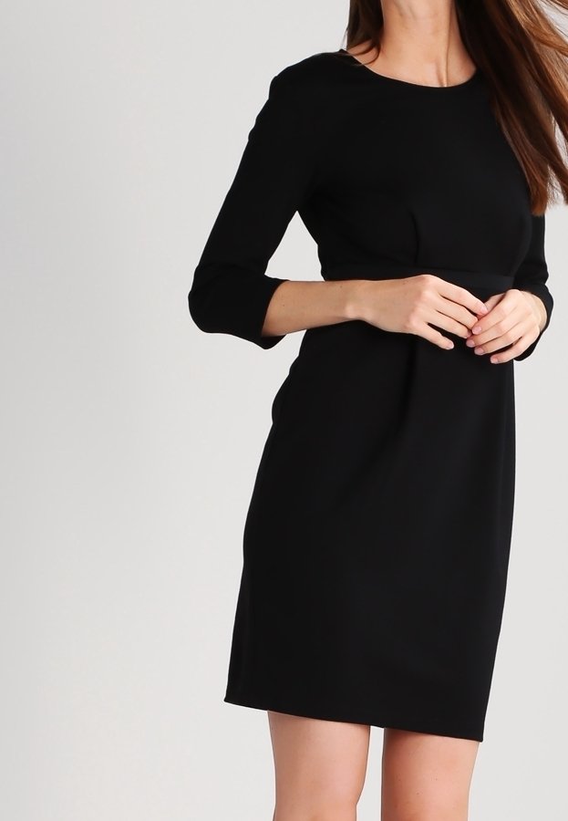 Femme portant une robe noire simple longueur genou avec des manches trois-quarts, debout devant un fond clair uni.