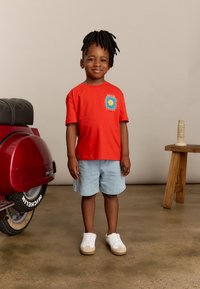 Rode katoenen T-shirt met blauwe grafische print, gecombineerd met lichte denimshorts. Witte sneakers met beige accenten. Achtergrond toont een vintage scooter.