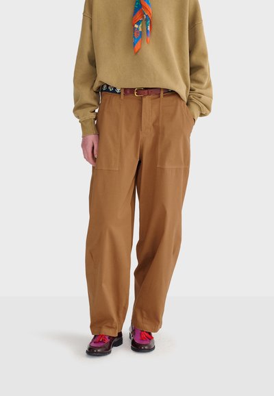 Personne portant un pantalon marron ample, une chemise beige à manches longues, une écharpe colorée et des chaussures foncées avec des pompons roses vifs, les mains dans les poches.