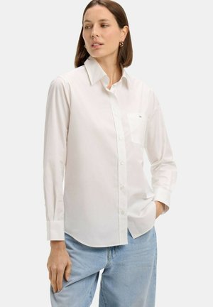 Femme portant une chemise blanche à manches longues avec boutons et poche, ainsi qu'un jean bleu clair, debout avec une main dans la poche, regardant sur le côté.