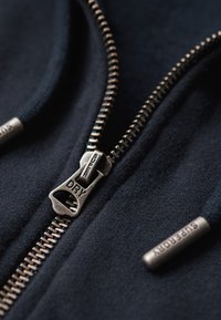 Granatowa bluza z kapturem na zamek błyskawiczny, z metalowym zamkiem i wygrawerowaną etykietą "DRY". Gładka struktura oraz regulowane sznurki ze wstawkami "SUPERDRY".