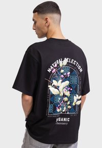 Schwarzes, oversize T-Shirt mit floralem Grafikdesign in Weiß, Blau und Pink auf der Rückseite, mit Textangaben "Natürliche Selektion" und "Organische Chemie."
