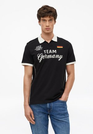 Młody mężczyzna w czarnej koszulce polo z białym kołnierzykiem i napisem "Team Germany", logo Mistrzostw Świata w Piłce Nożnej 2026 oraz naszywką z niemiecką flagą.