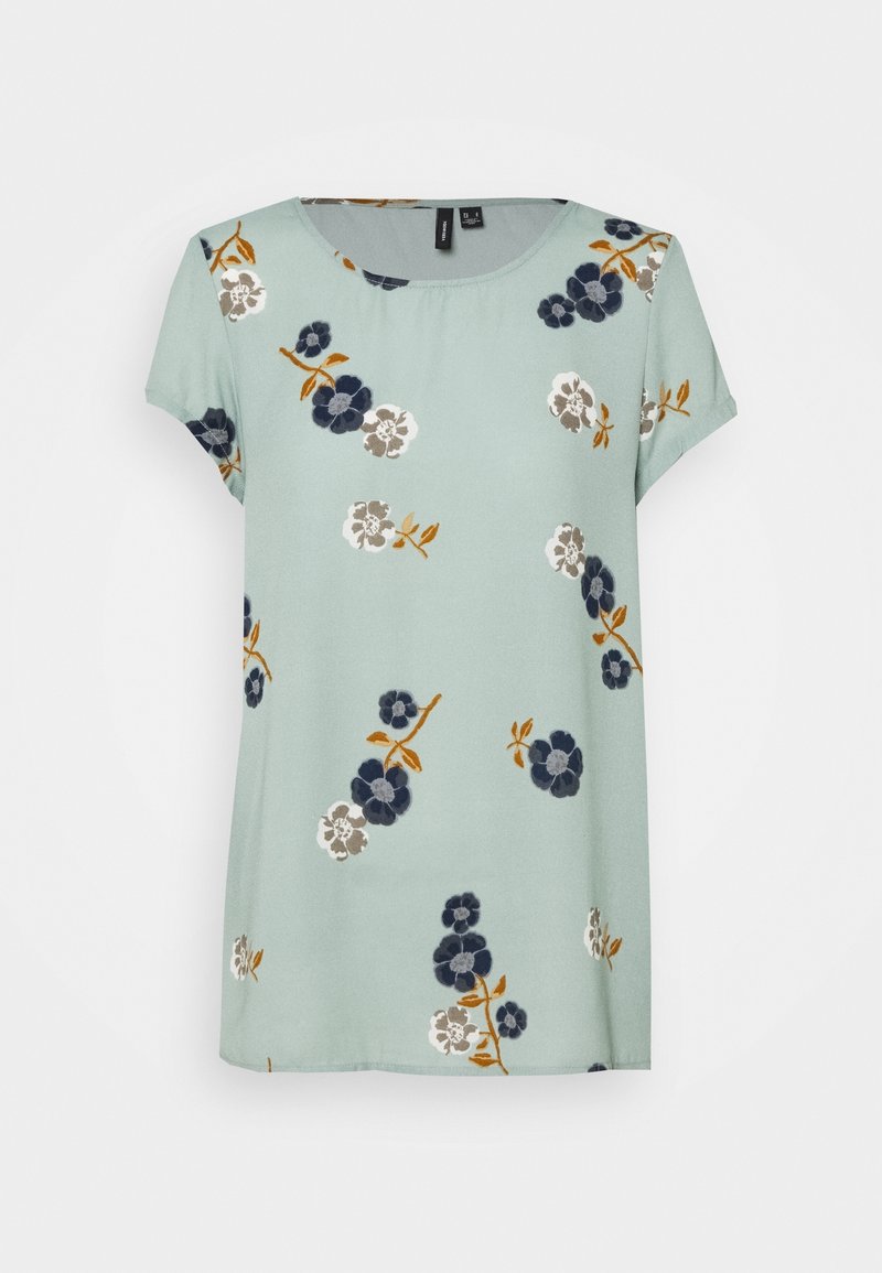 Blouse bleu clair à manches courtes avec col rond, décorée de motifs floraux dispersés bleu marine, blanc et beige avec des tiges couleur moutarde.
