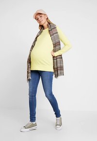 Mulher grávida vestindo um suéter amarelo, jeans azuis, cachecol xadrez, boné bege e ténis verdes, parada com uma mão no bolso.