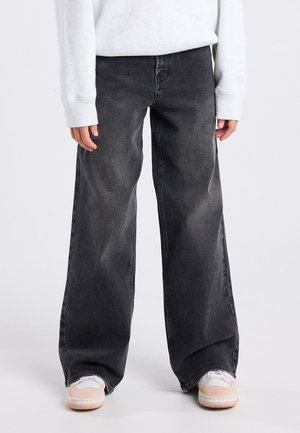 Person, der bærer falmede sorte vidde jeans, hvid sweatshirt og hvide sneakers med lyserøde detaljer, står mod en ensfarvet baggrund.
