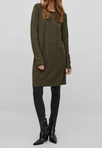 Abito maglione color verde oliva con maniche lunghe, polsini a coste e spacchi laterali. Indossato sopra jeans skinny neri e stivaletti neri con punta.